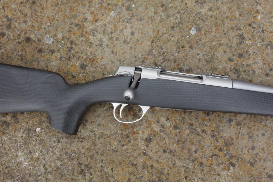Sako 90 Peak Carbon rifle with carbon fibre stock, fluted barrel, adjustable trigger, and detachable magazine, showcased on a textured surface.