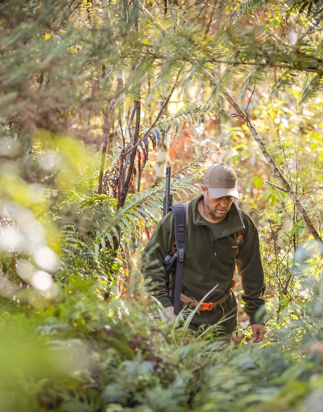 Swazi Doughroaster Fleece Jumper in use: Man walking in woods, wearing a hat, green jacket, and backpack, highlighting its outdoor functionality and comfort.