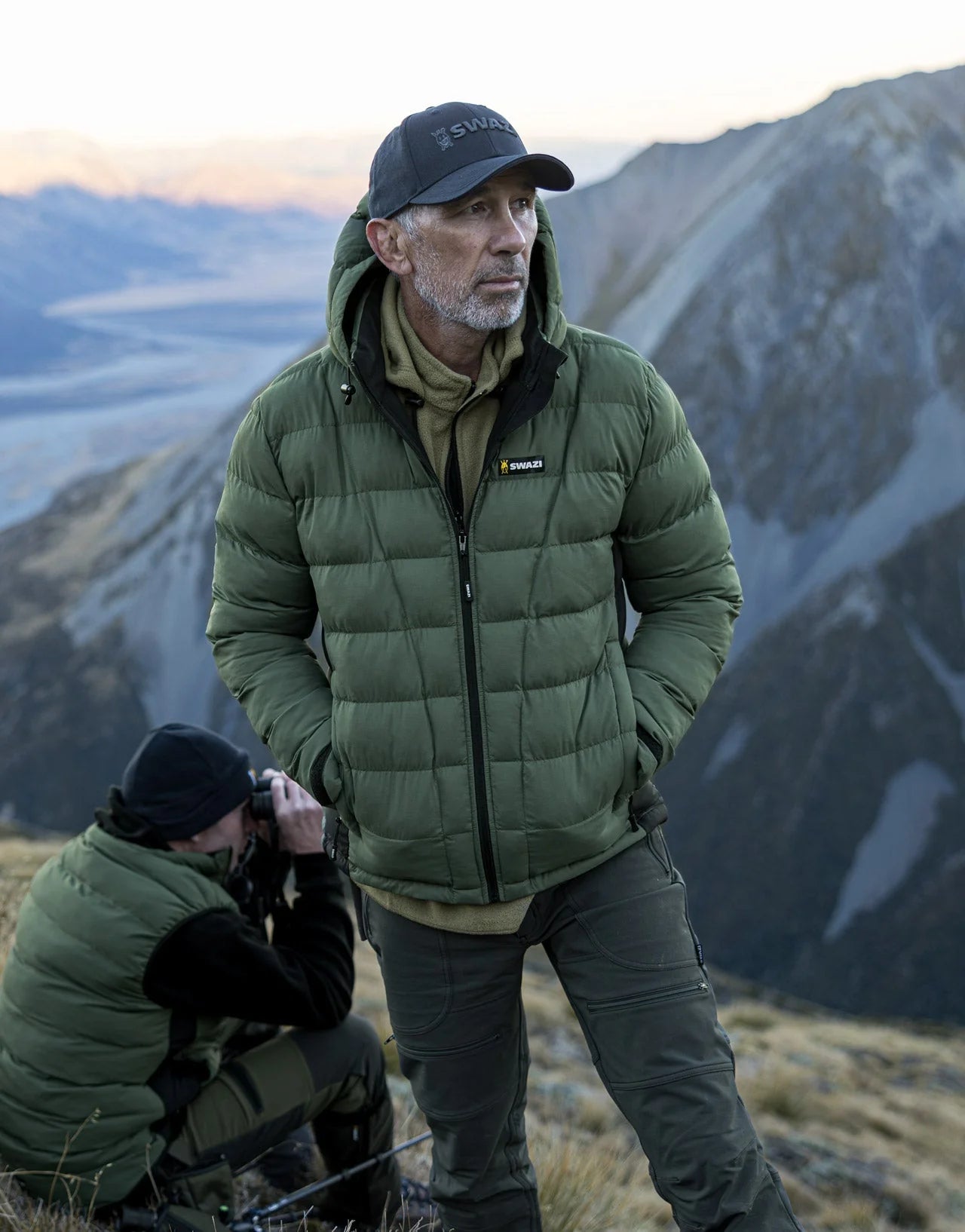 Swazi Puffadder Men's Puffer Jacket with detachable hood, shown on a mountaineer standing outdoors, highlighting its tailored fit and practical features for cold conditions.