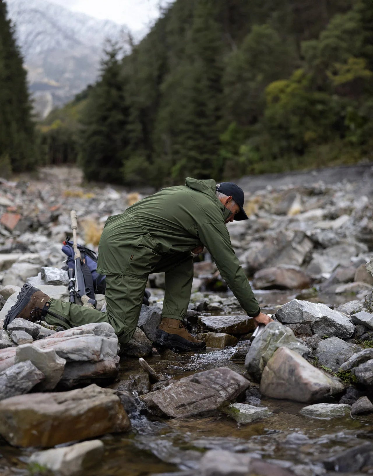 Man in a Swazi Tahr Ultralite Jacket kneels on rocky riverbed, highlighting its lightweight, waterproof design ideal for hiking and outdoor adventures.