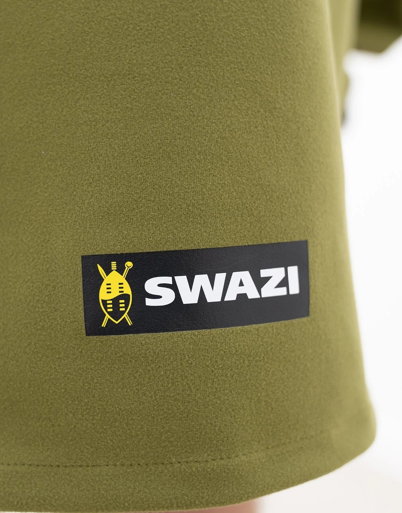Swazi Kagoule Anorak, a quiet waterproof hunting jacket, showcases a close-up of its green fabric and distinctive logo, highlighting its durable, water-resistant features.