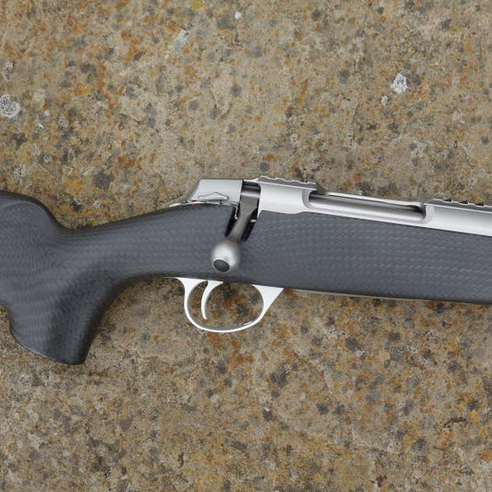 Sako 90 Peak Carbon rifle with carbon fibre stock, fluted barrel, adjustable trigger, and detachable magazine, showcased on a textured surface.