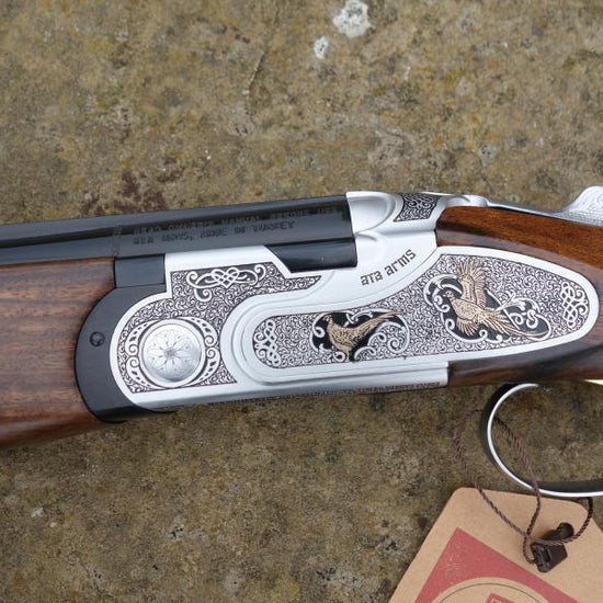 Close-up of the ATA Elegant Gold Game gun, showcasing its handle and trigger, with a brown tag attached. Features include multi chokes and automatic safety catch.