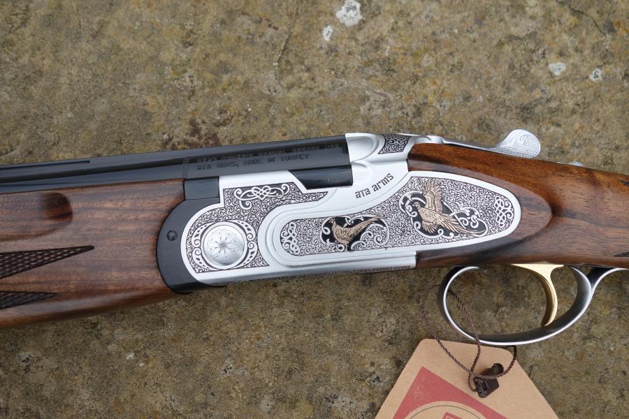 Close-up of the ATA Elegant Gold Game gun, showcasing its handle and trigger, with a brown tag attached. Features include multi chokes and automatic safety catch.