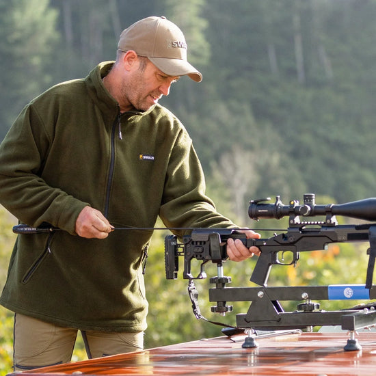 Man wearing Swazi Doughroaster Fleece Jumper, holding a rifle outdoors. Jumper features high collar, long chest zip, and kangaroo pocket for functionality.