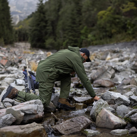 Man in a Swazi Tahr Ultralite Jacket kneels on rocky riverbed, highlighting its lightweight, waterproof design ideal for hiking and outdoor adventures.